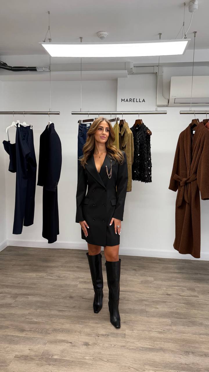 Camilla Brossa a figura intera nello showroom Marella, in abito blazer nero doppiopetto con catena dorata e stivali al ginocchio, davanti agli stender