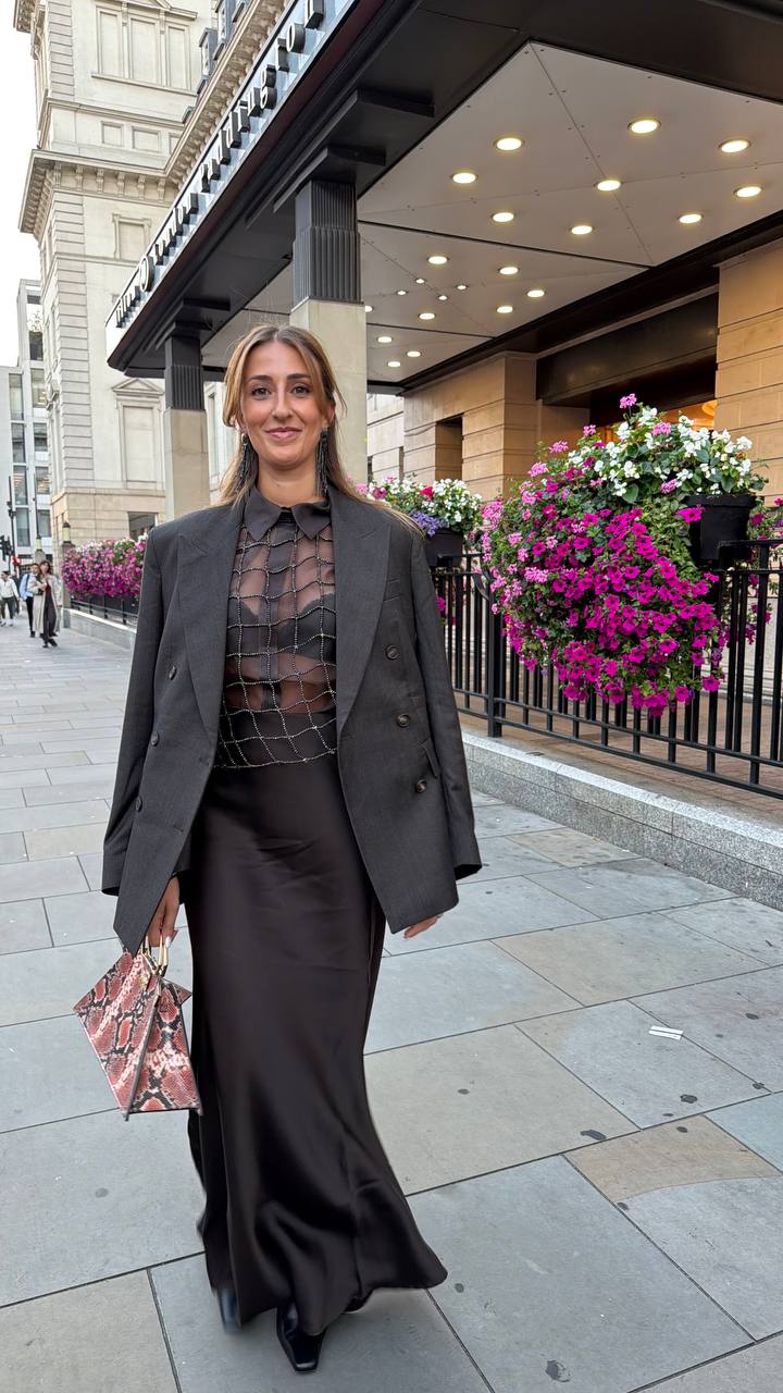 Camilla Brossa cammina per Londra davanti all’ingresso di un hotel fiorito, in blazer grigio oversize, top di cristalli e gonna lunga in raso, con borsa stampa pitone