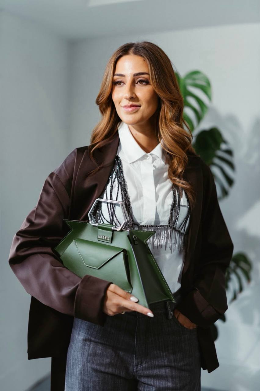 Camilla Brossa in giacca di raso color cioccolato sopra la camicia bianca, con una borsa verde dal manico esagonale sotto il braccio