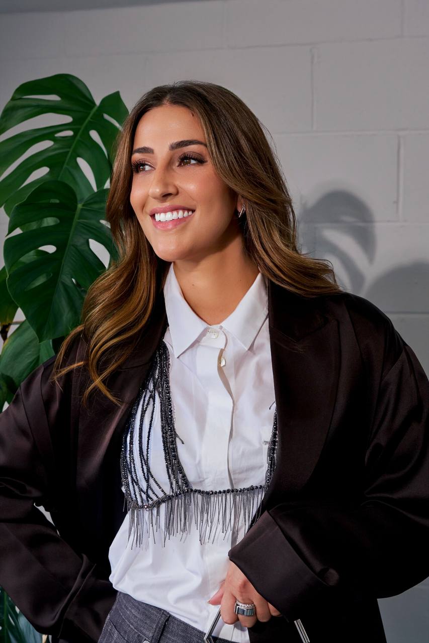 Primo piano di Camilla Brossa sorridente, in giacca di raso e bralette a frange di cristalli, accanto a una monstera